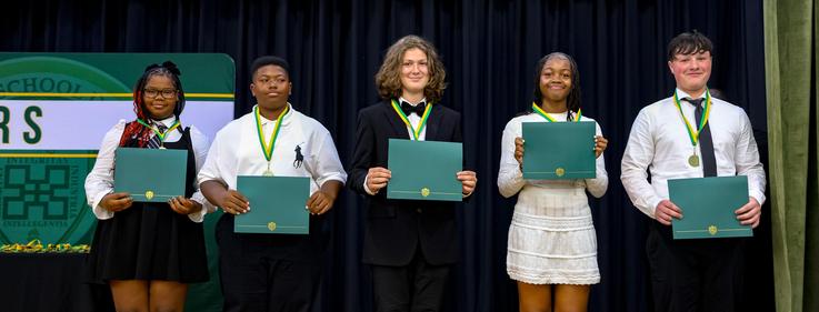 Five graduates in formal attire holding certificates on stage, smiling at the camera.