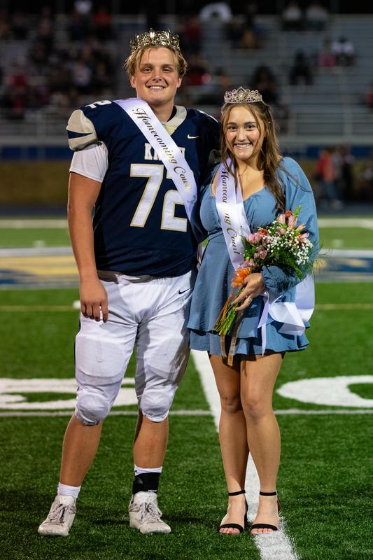 boy in a football uniform and a crown poses with a girl in a dress holding flowers and wearing a crown