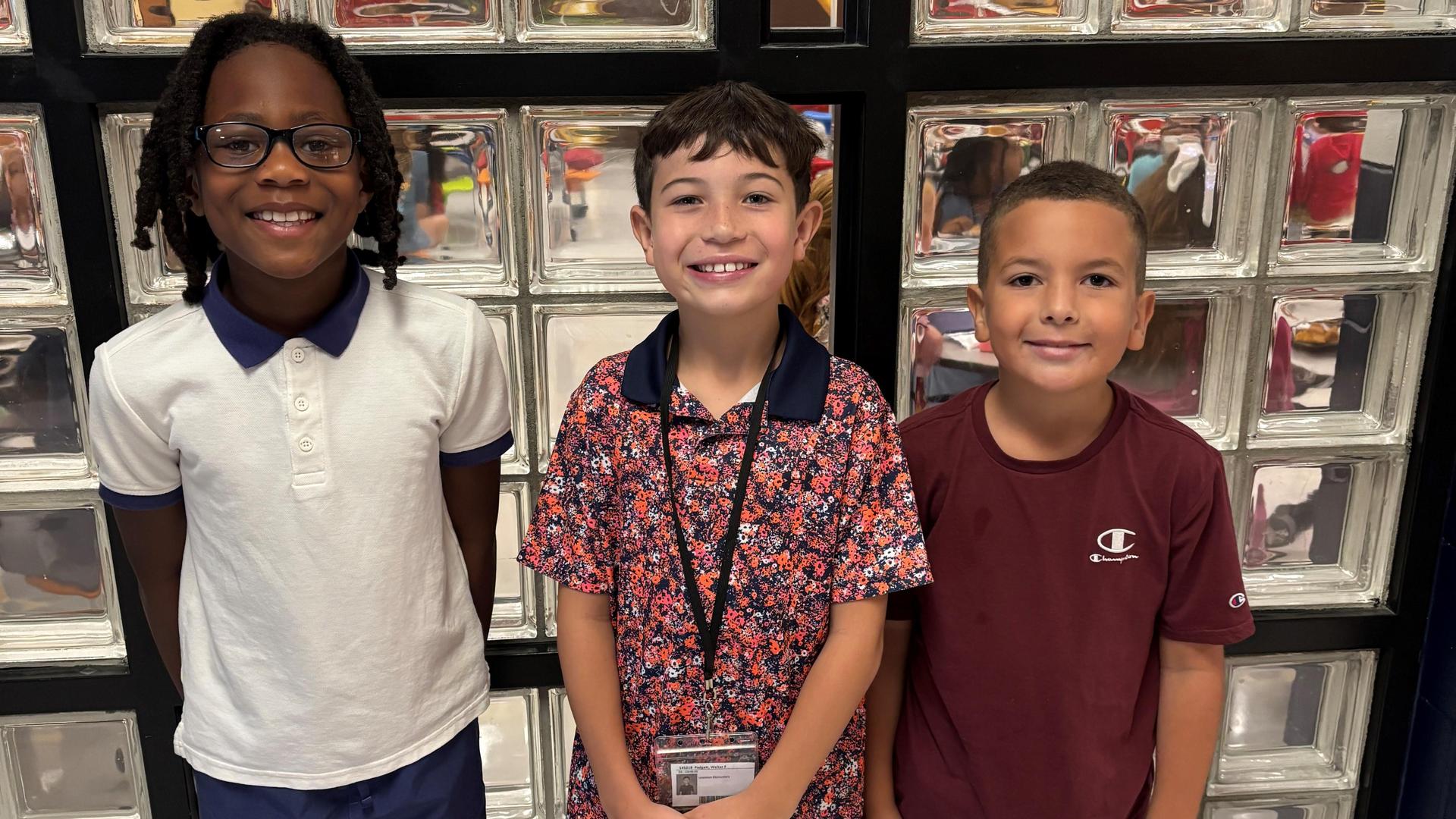 Three smiling boys standing together in front of a glass block wall.