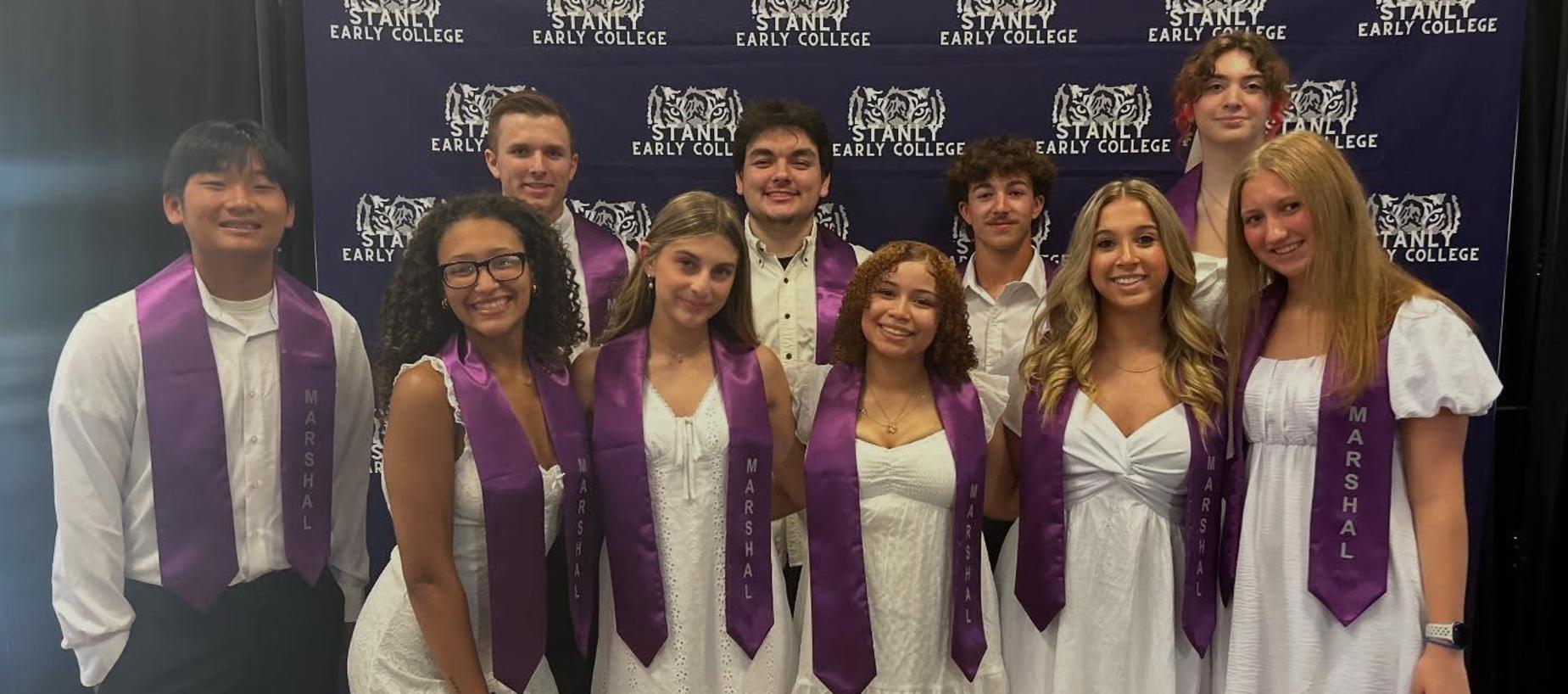 Group of students in white attire with purple sashes, posing in front of a backdrop.