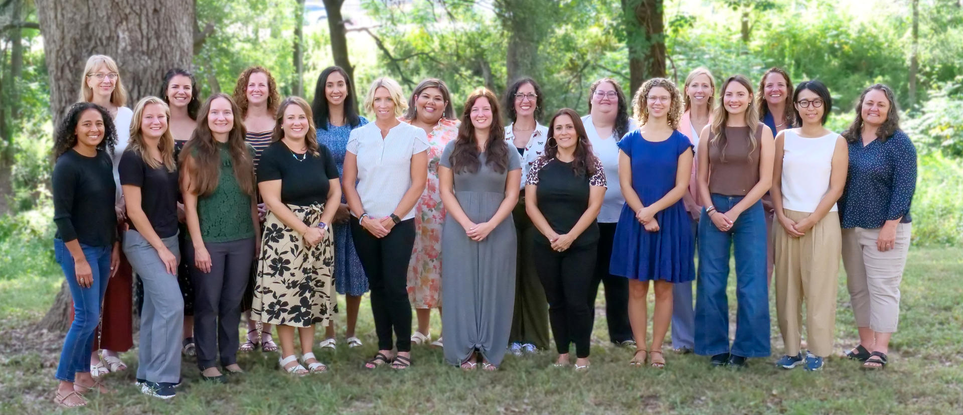Group photo of 17 women standing outside in front of a brick building, smiling on a sunny day.