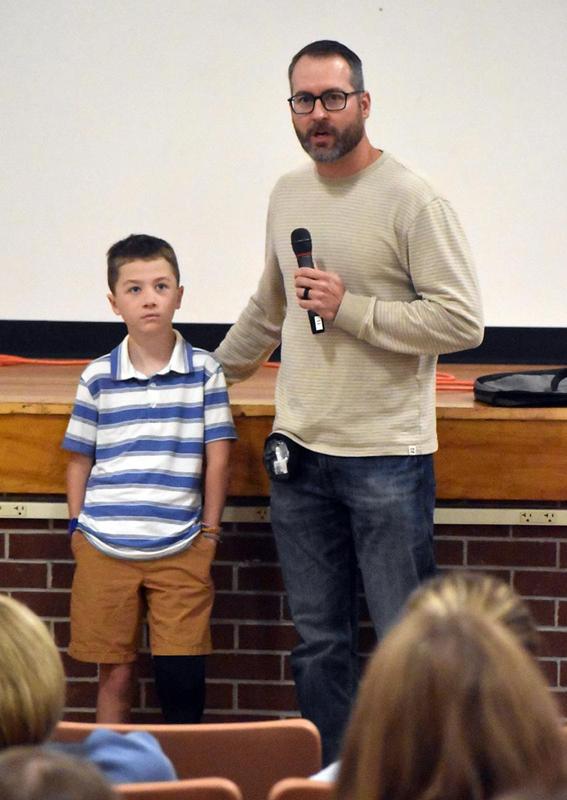 A man holds a microphone next to a boy, both speaking to an audience in a classroom.