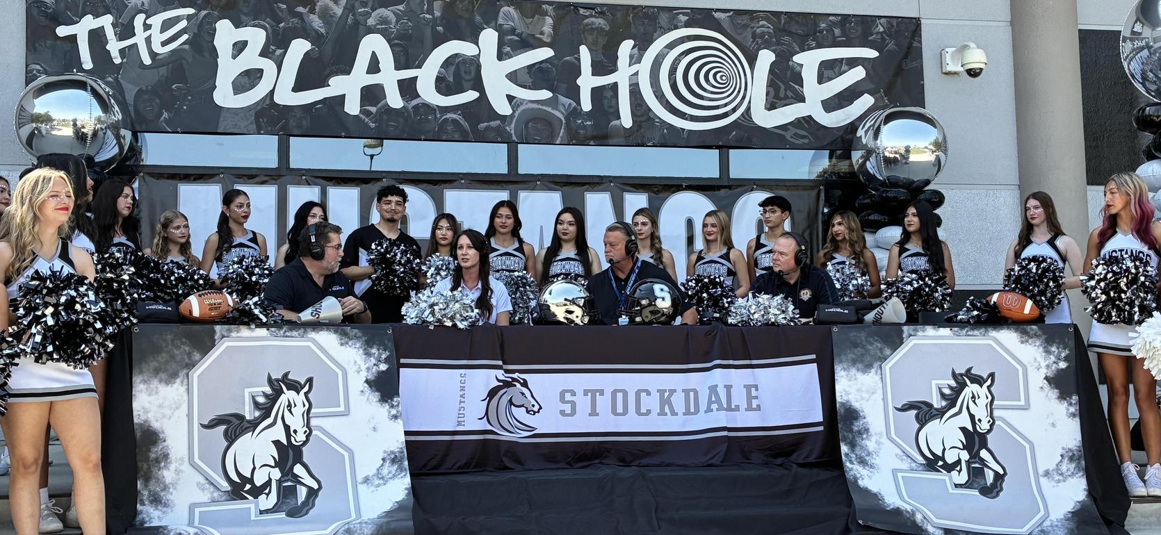 A cheerleading squad and announcers gather at a sports event under a large banner.