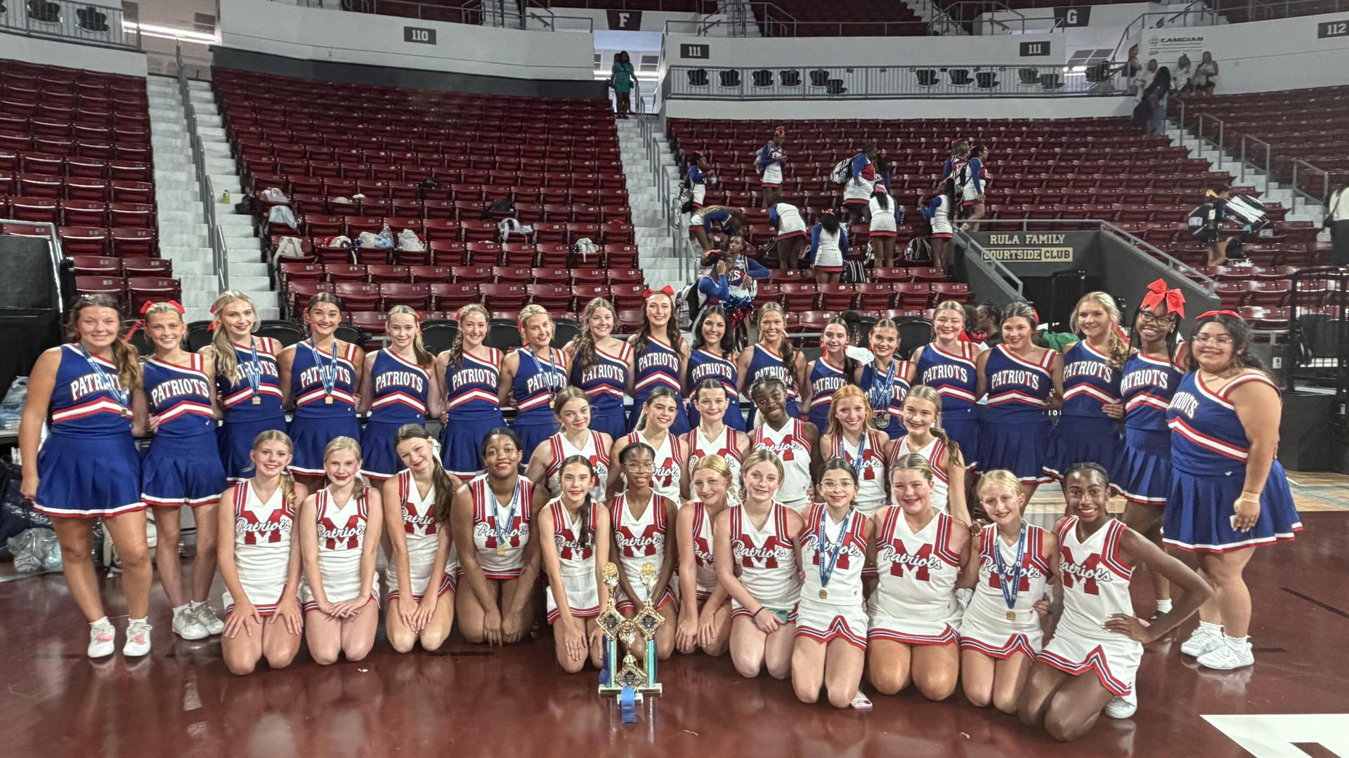 Large group of cheerleaders posing with a trophy in a gym.