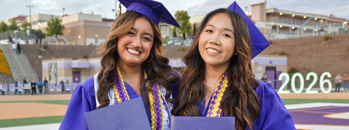 Two graduates in purple gowns holding diplomas, smiling with a stadium backdrop.