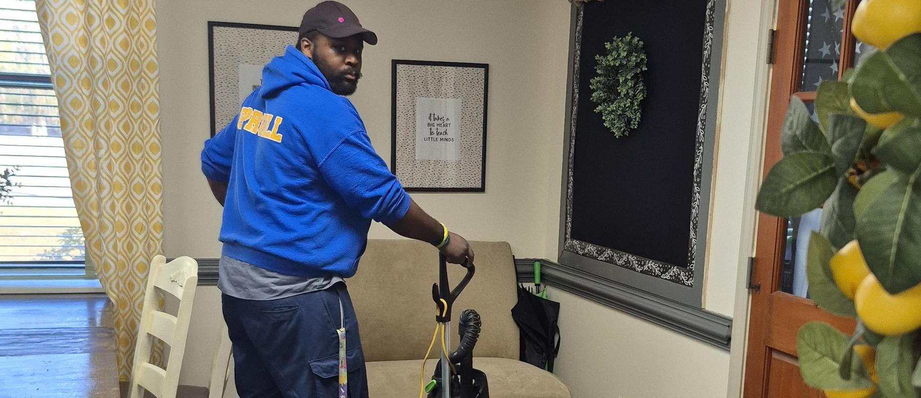 A person vacuuming a room with a carpet cleaner, wearing blue attire.