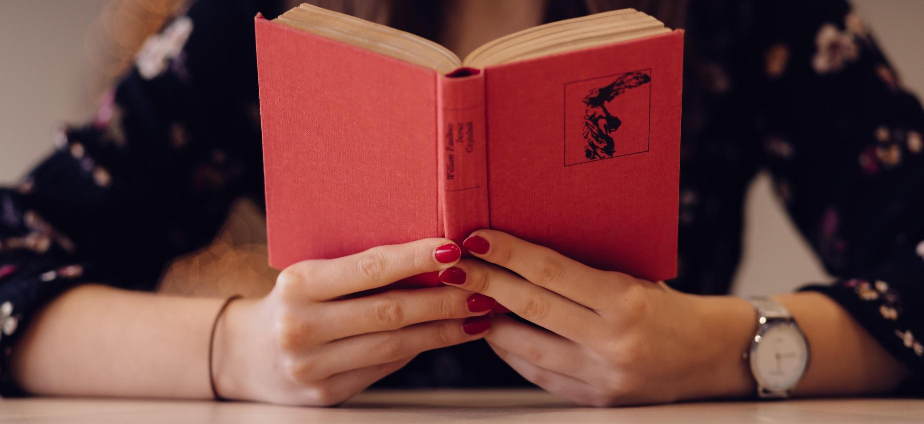 A person with red nails holds a red book open on a table.