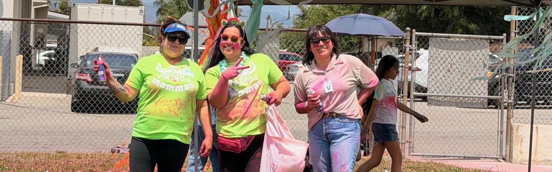 Three smiling women in colorful shirts, posing under a tent with playful expressions.