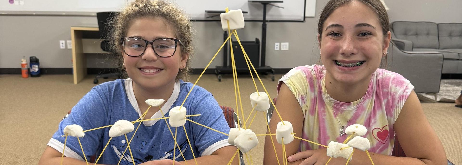 Two girls smile beside a structure made of spaghetti and marshmallows.