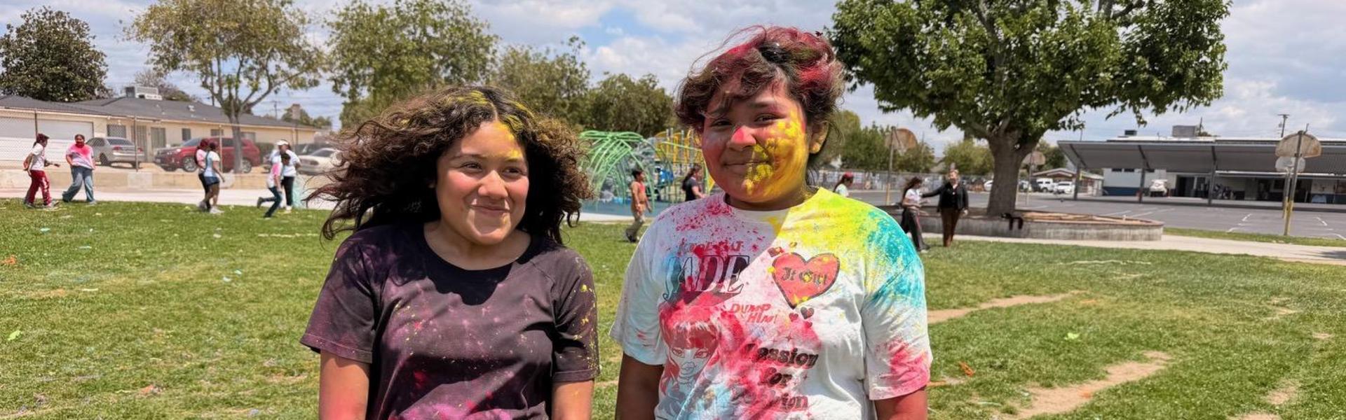 Two kids in colorful shirts, smiling brightly against a grassy background.