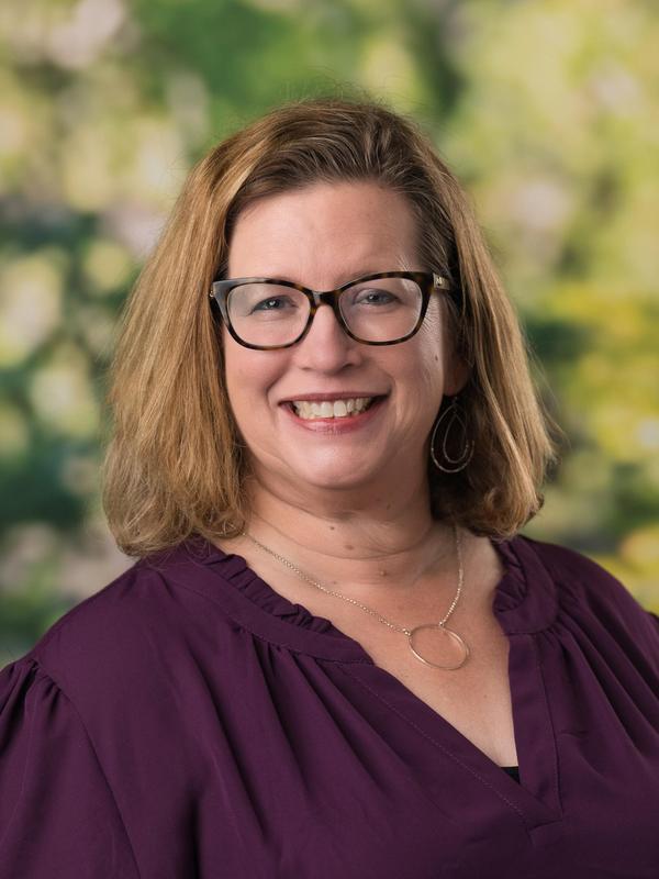 Smiling woman with glasses and light brown hair, wearing a purple blouse.