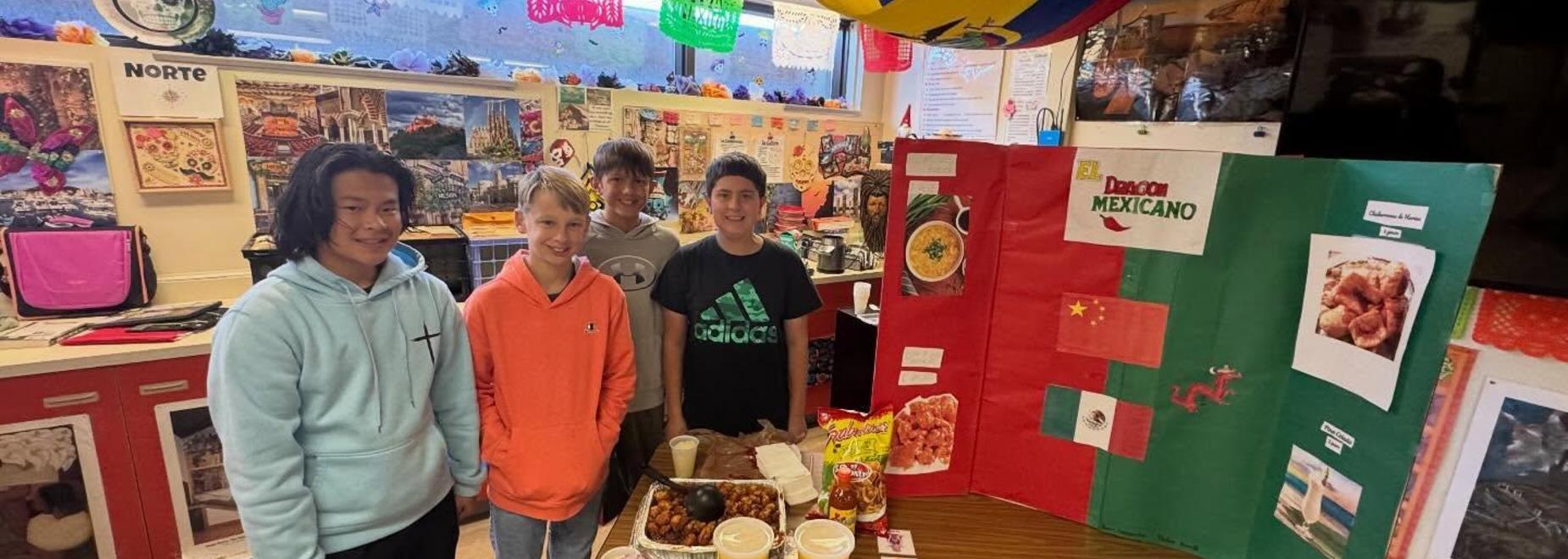 Four boys stand around a food display with a tri-fold poster in a vibrant classroom setting.