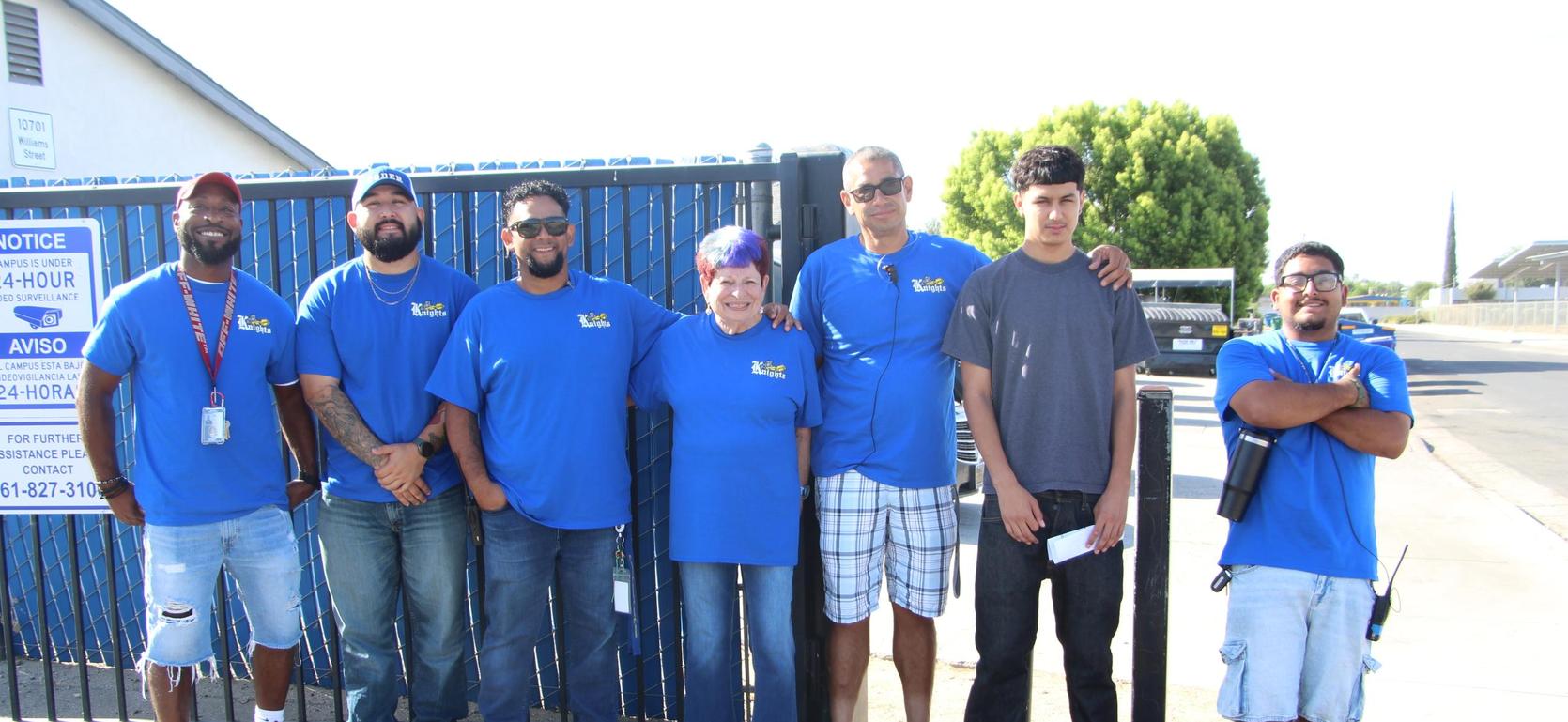 Seven people in blue shirts stand together by a gate, smiling at the camera.