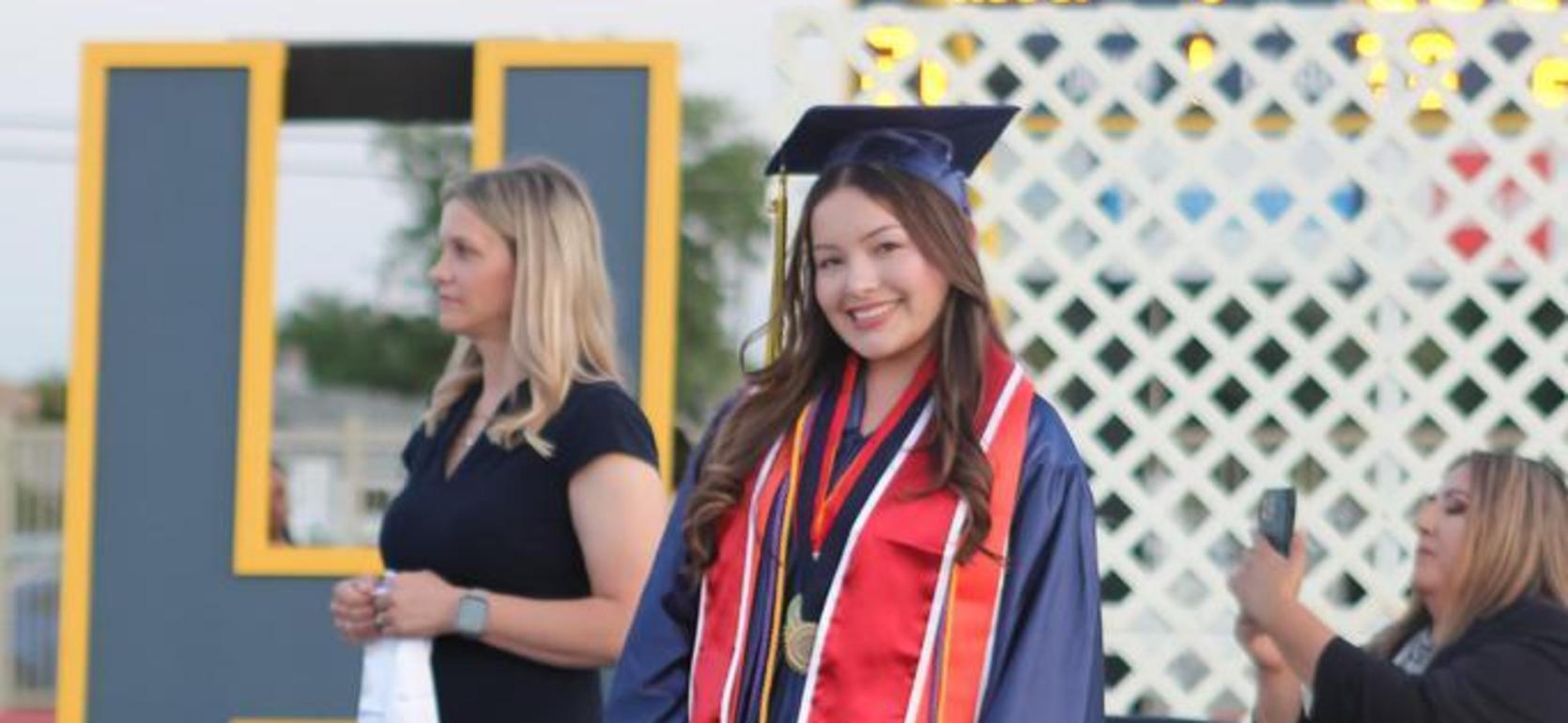 Graduate in cap and gown with medals, smiling at a ceremony with attendees behind her.