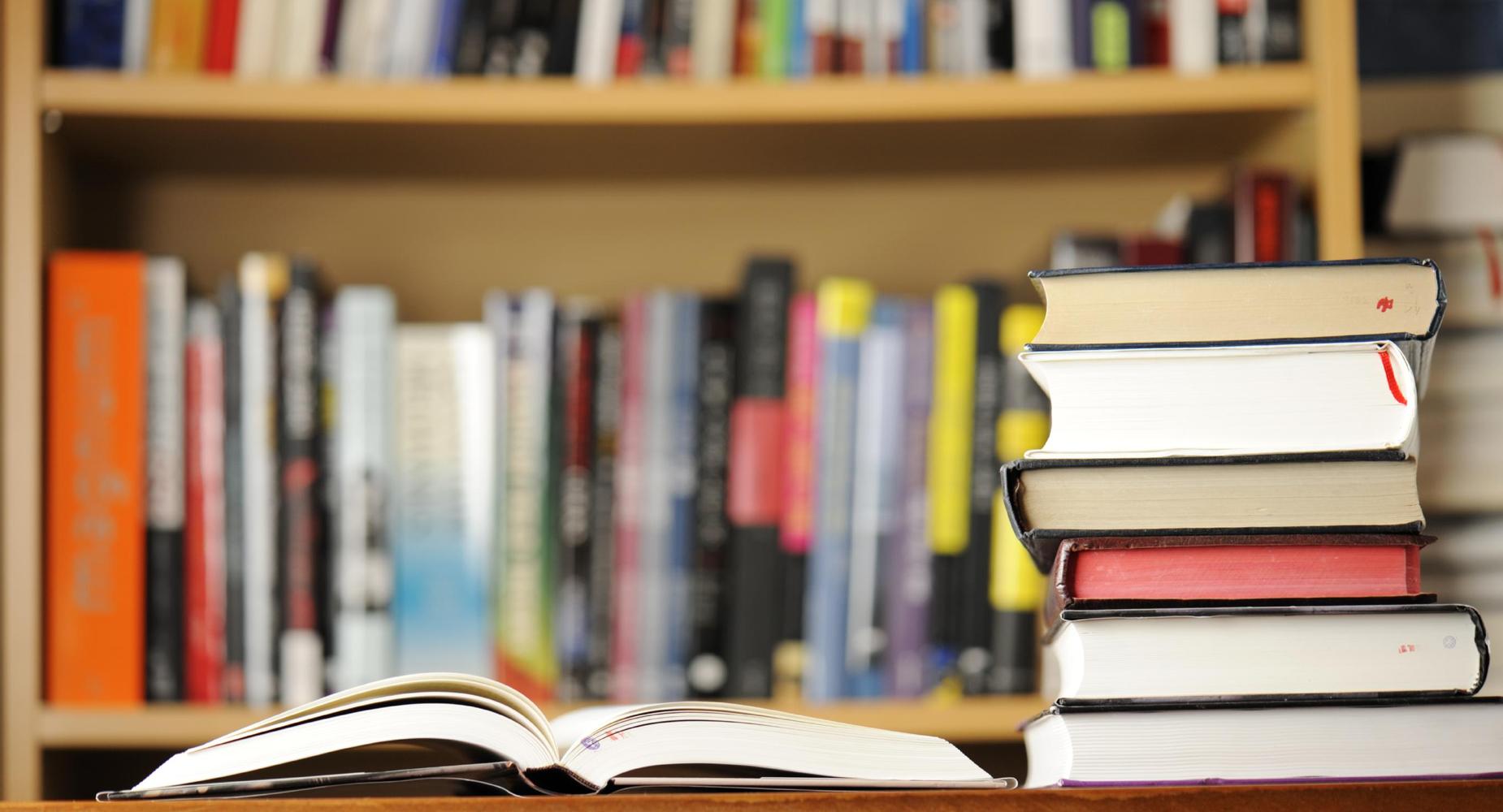 An open book in the foreground with stacked books and a blurred bookshelf in the background.