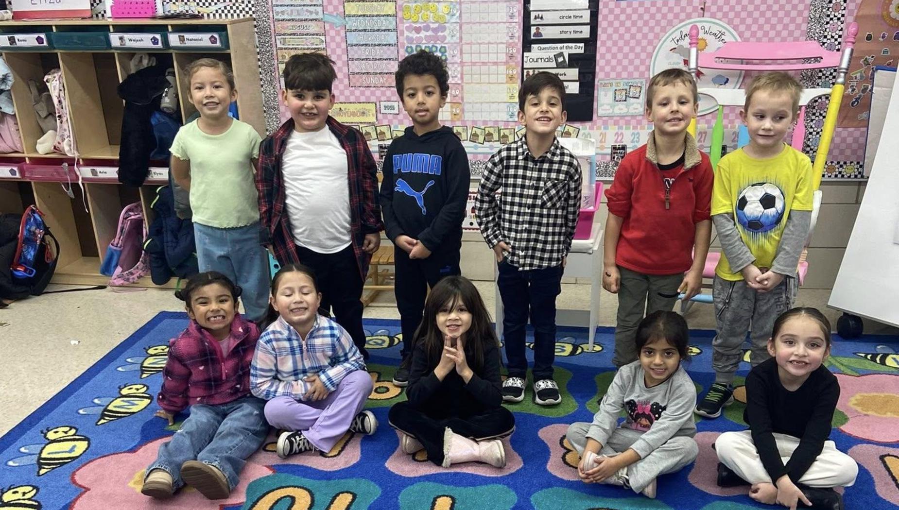 Group of children sitting on a colorful rug with letters, smiling and posing for the camera.