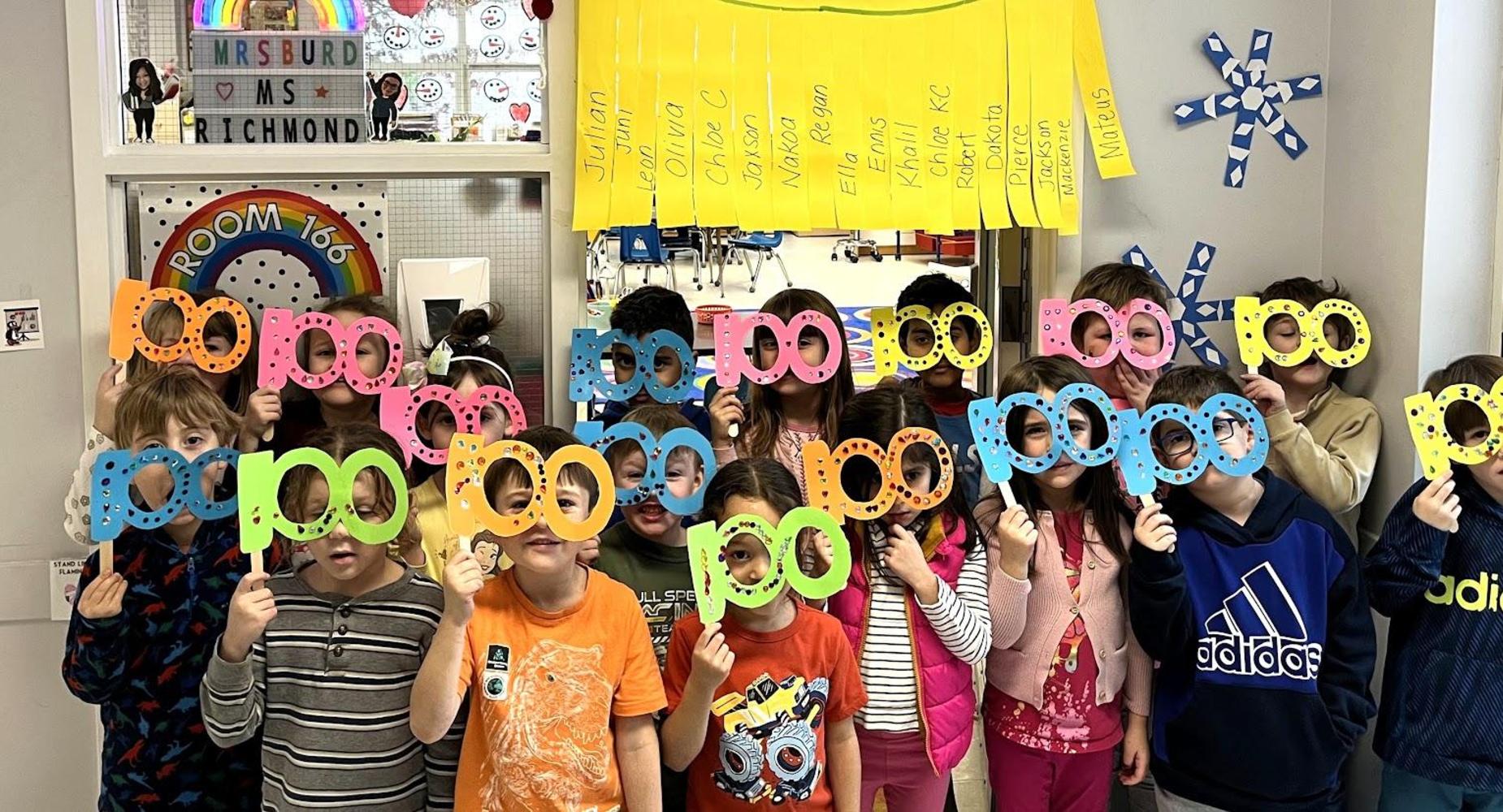 Students hold colorful paper glasses in front of a classroom backdrop.