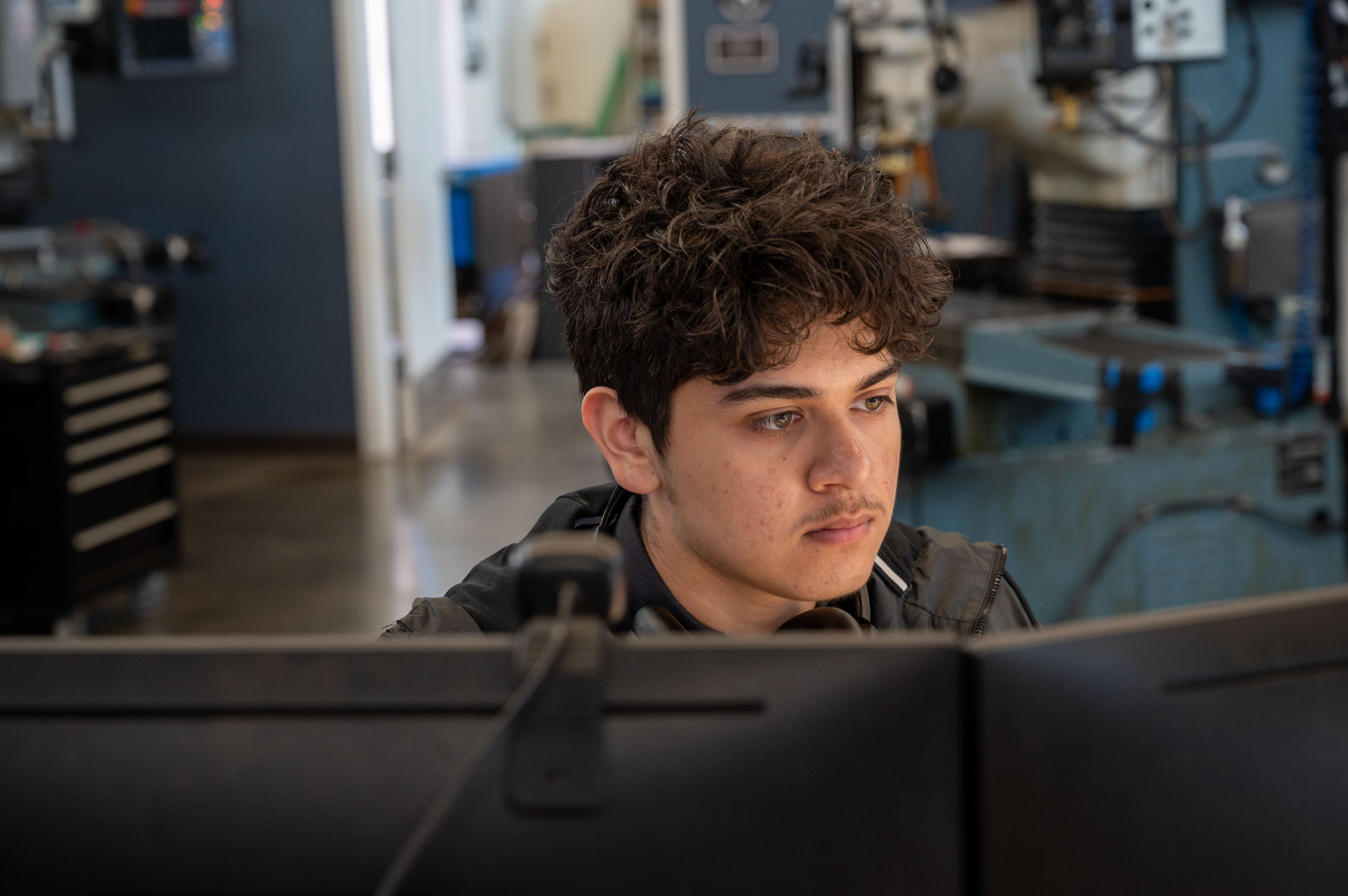 Student working at a computer in a technical classroom or lab, with industrial equipment and machinery visible in the background.