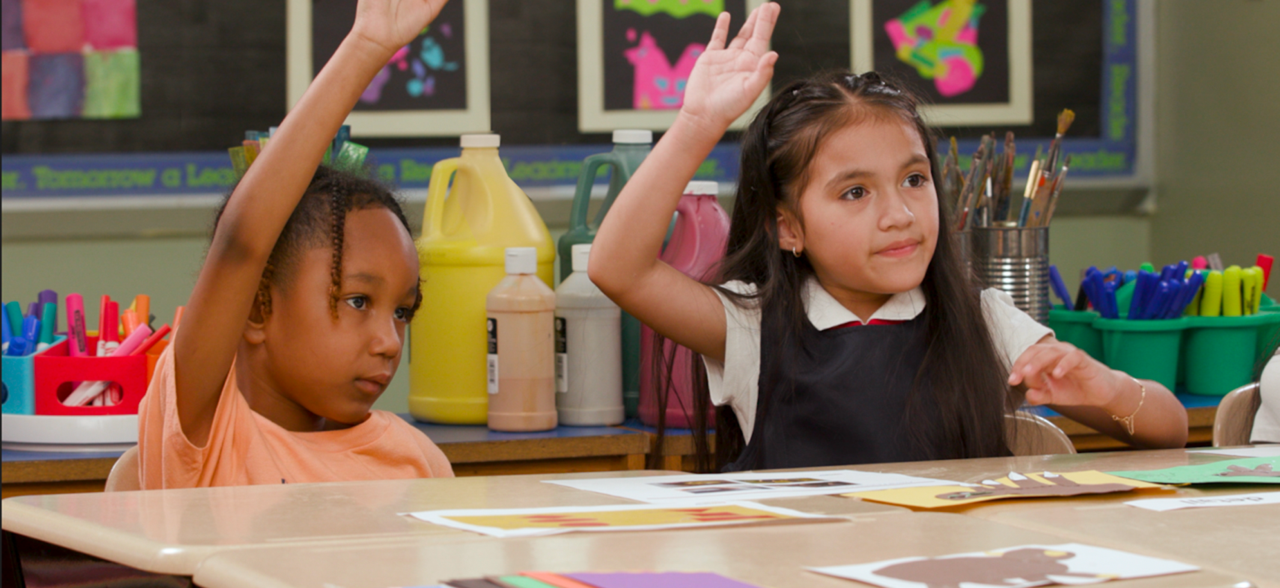 Two children in a classroom, one raising a hand and the other sitting quietly.