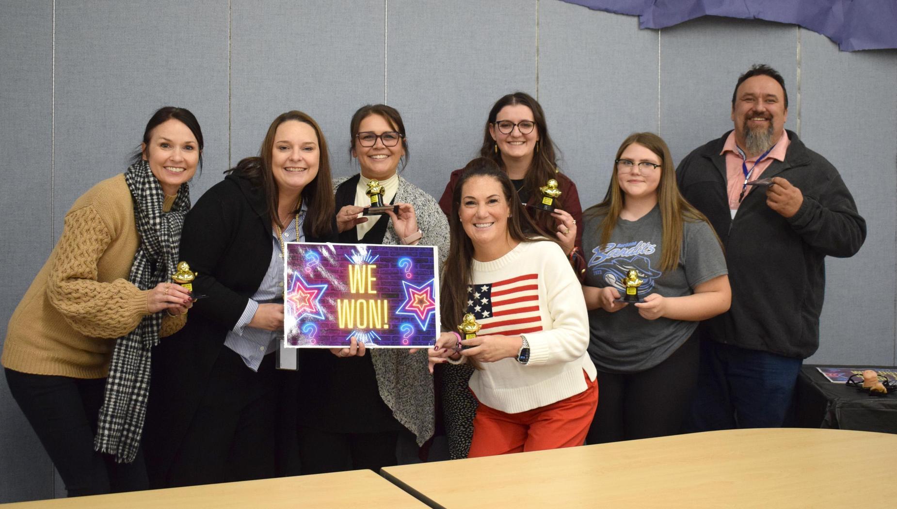 A team of smiling individuals holding trophies and celebrating a victory with a sign.