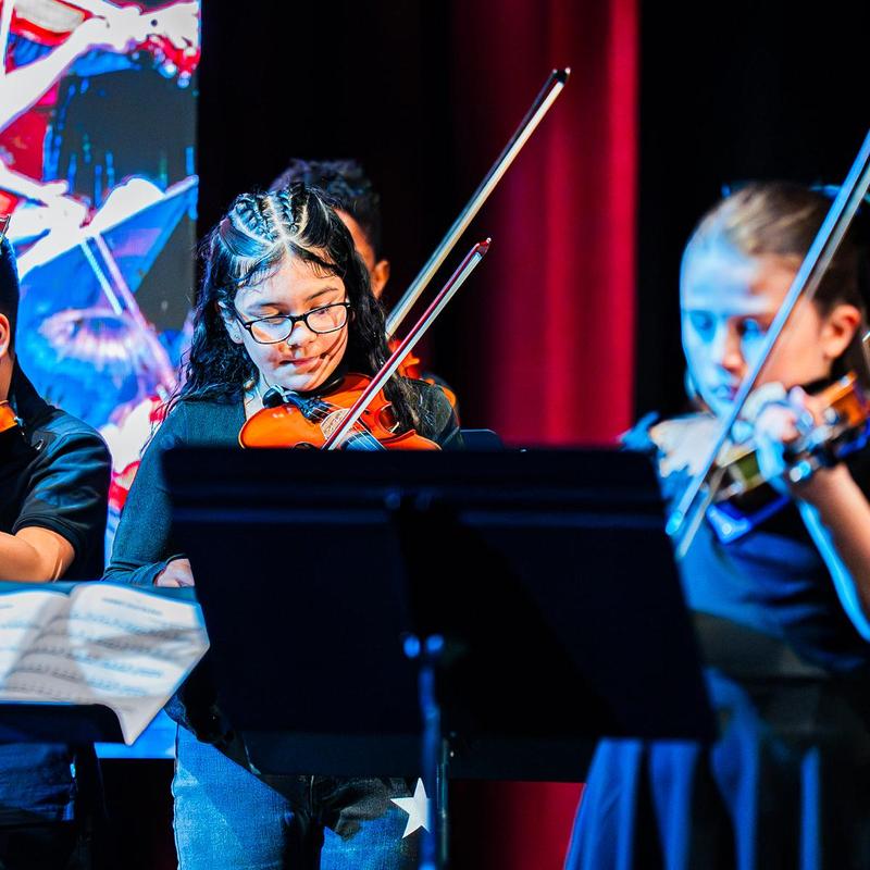 Young musicians playing violins on a stage with red curtains.
