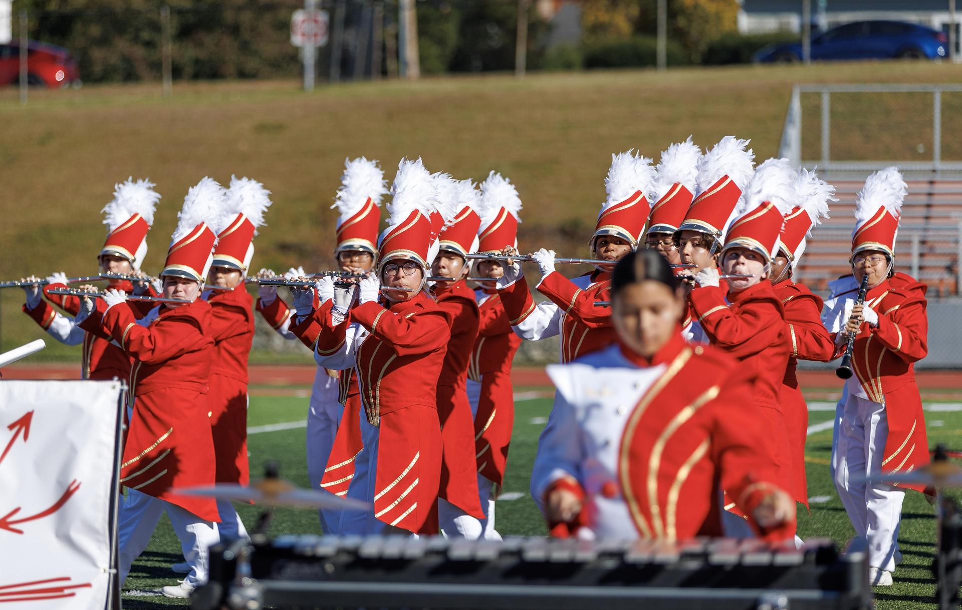 Marching band members in red uniforms performing with instruments outdoors.