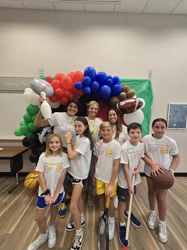 Smiling group of children posing with sports equipment in front of a balloon arch.
