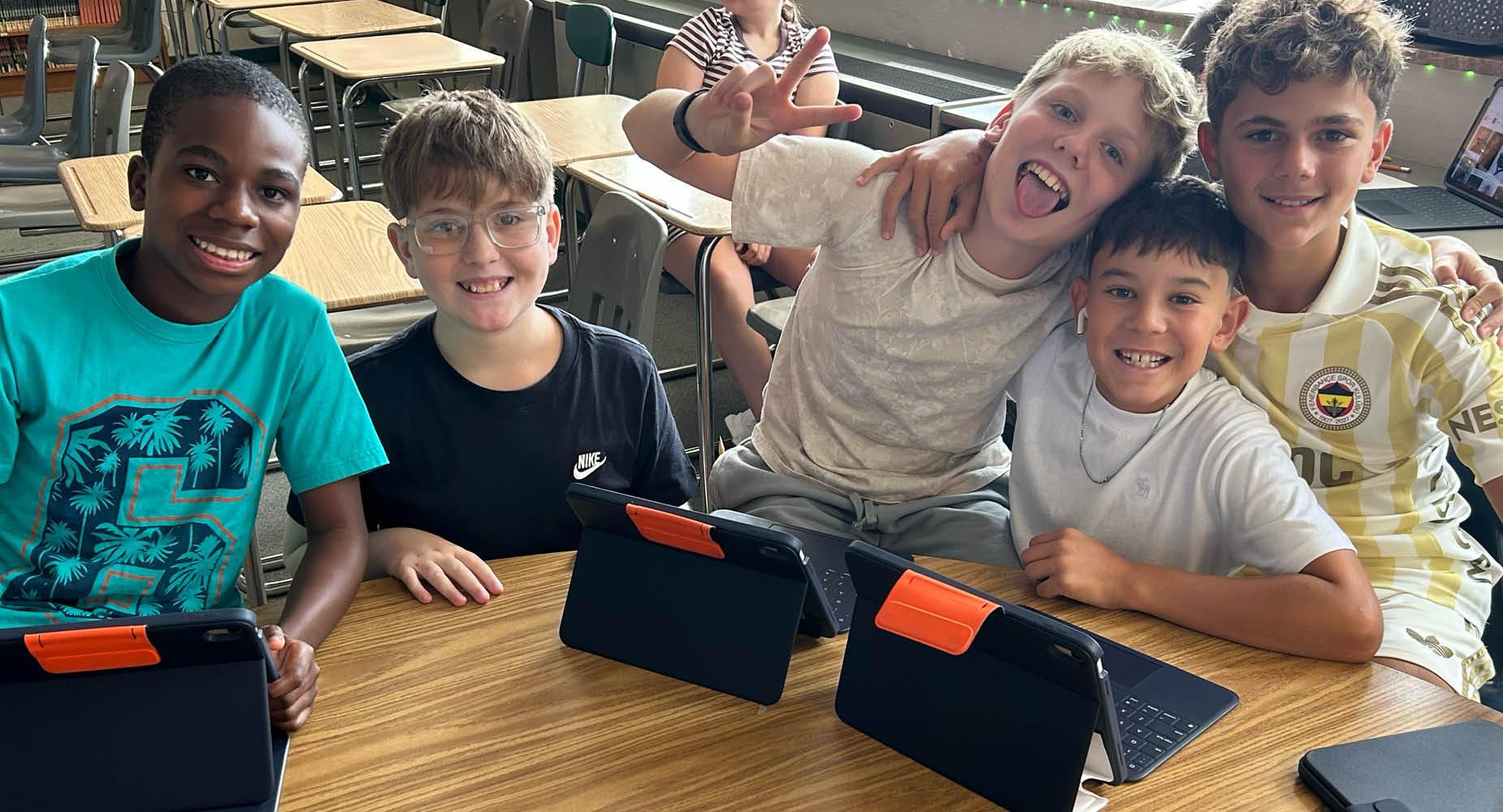 Five children sitting at a table, smiling and posing with tablets.