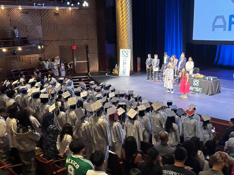 Graduates in silver gowns celebrate at a graduation ceremony on stage.