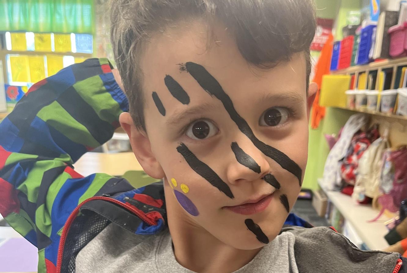 Child with painted face featuring black stripes and colorful dots in a classroom.