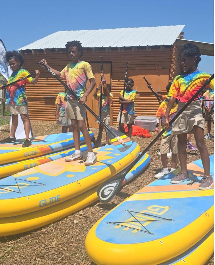 Scholars stand on paddle boards on grass with oars, learning how to steer