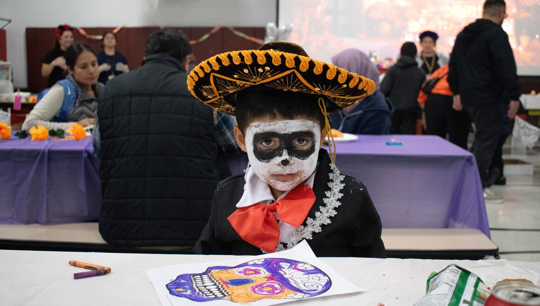 Child in a sombrero and face paint, coloring a skull drawing at a table.