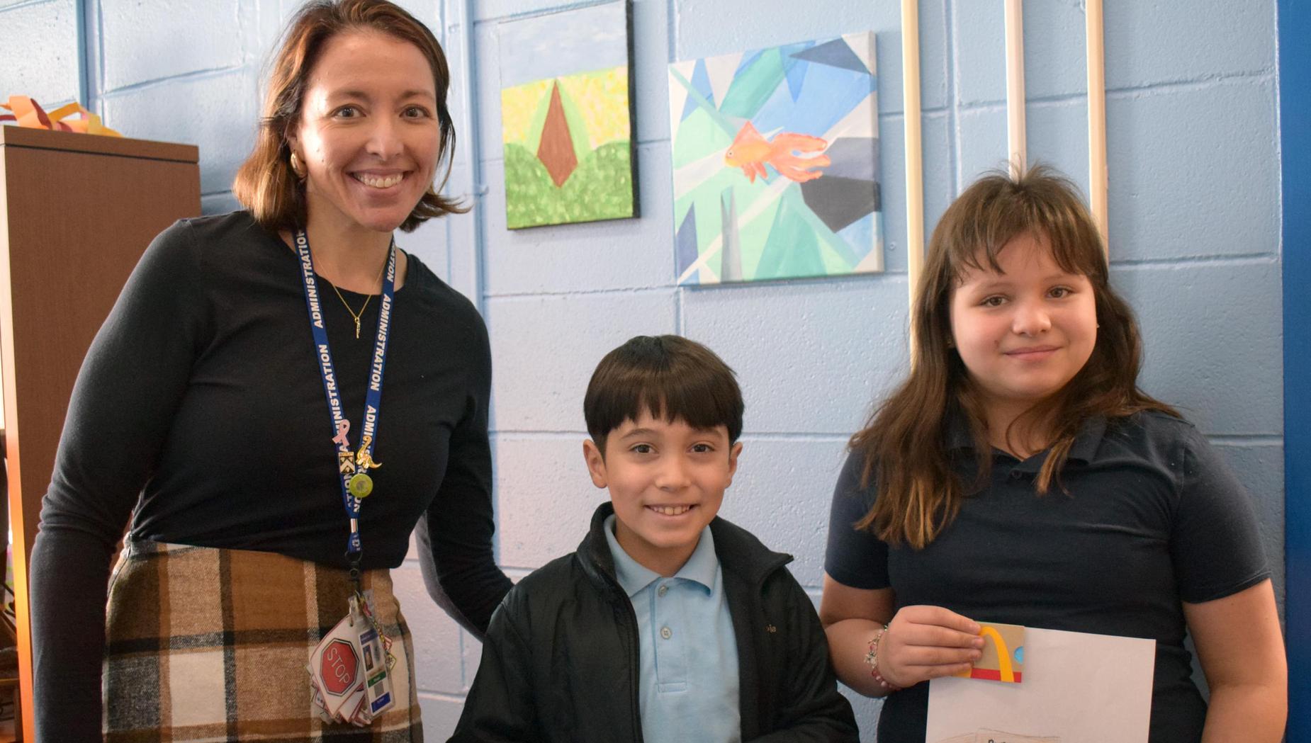 A teacher and two children smiling while holding artwork with a colorful letter design.