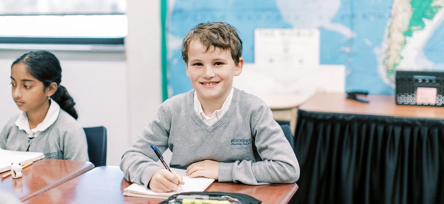 Smiling elementary student writes at his desk in front of a classroom map