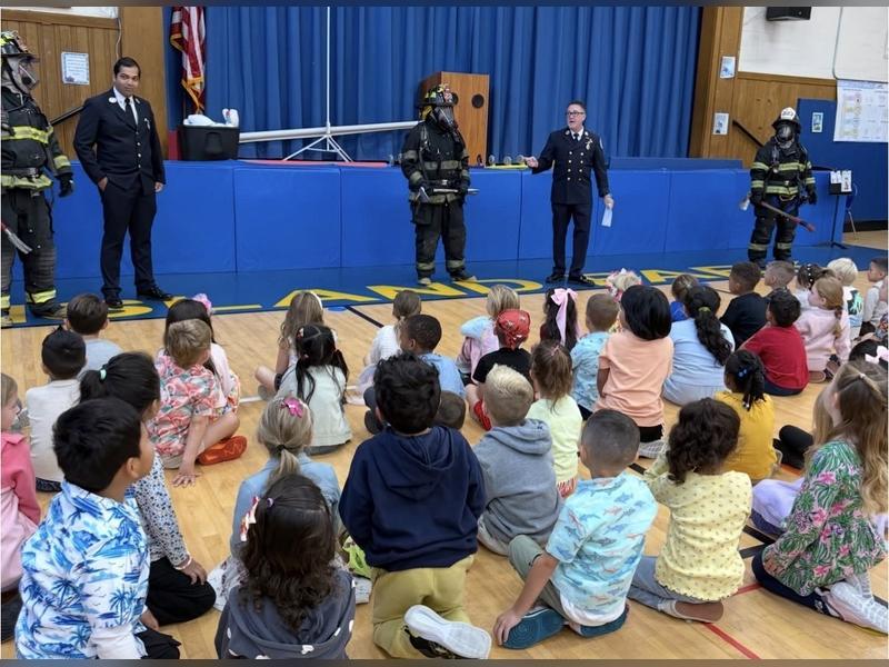 Firefighters in gear present safety tips to children seated on a gymnasium floor.