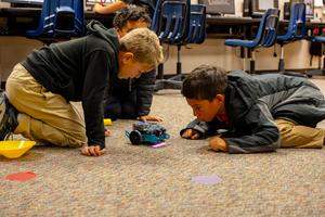Three children observe a remote-controlled car on the carpet.
