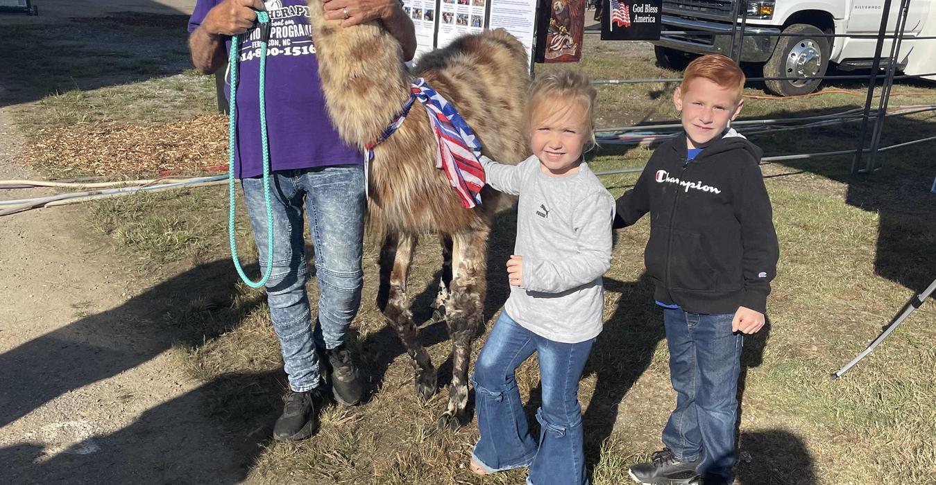 Two children posing with a goat wearing a patriotic ribbon at an outdoor fair setting.