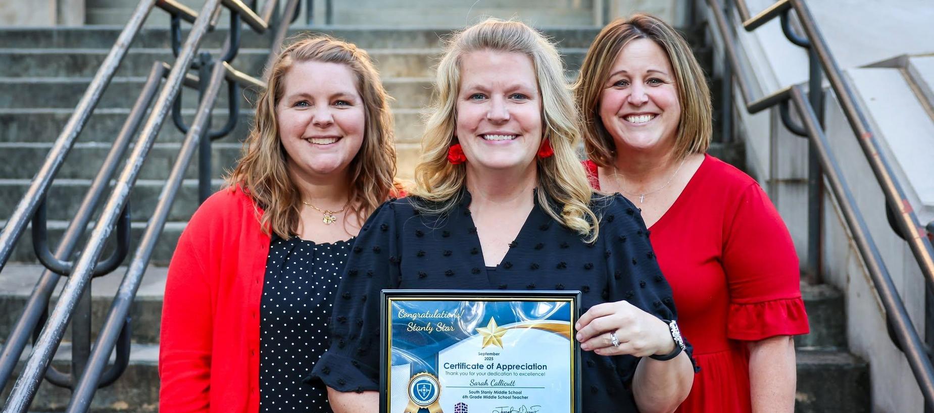 Three women holding a certificate of appreciation in front of stairs.