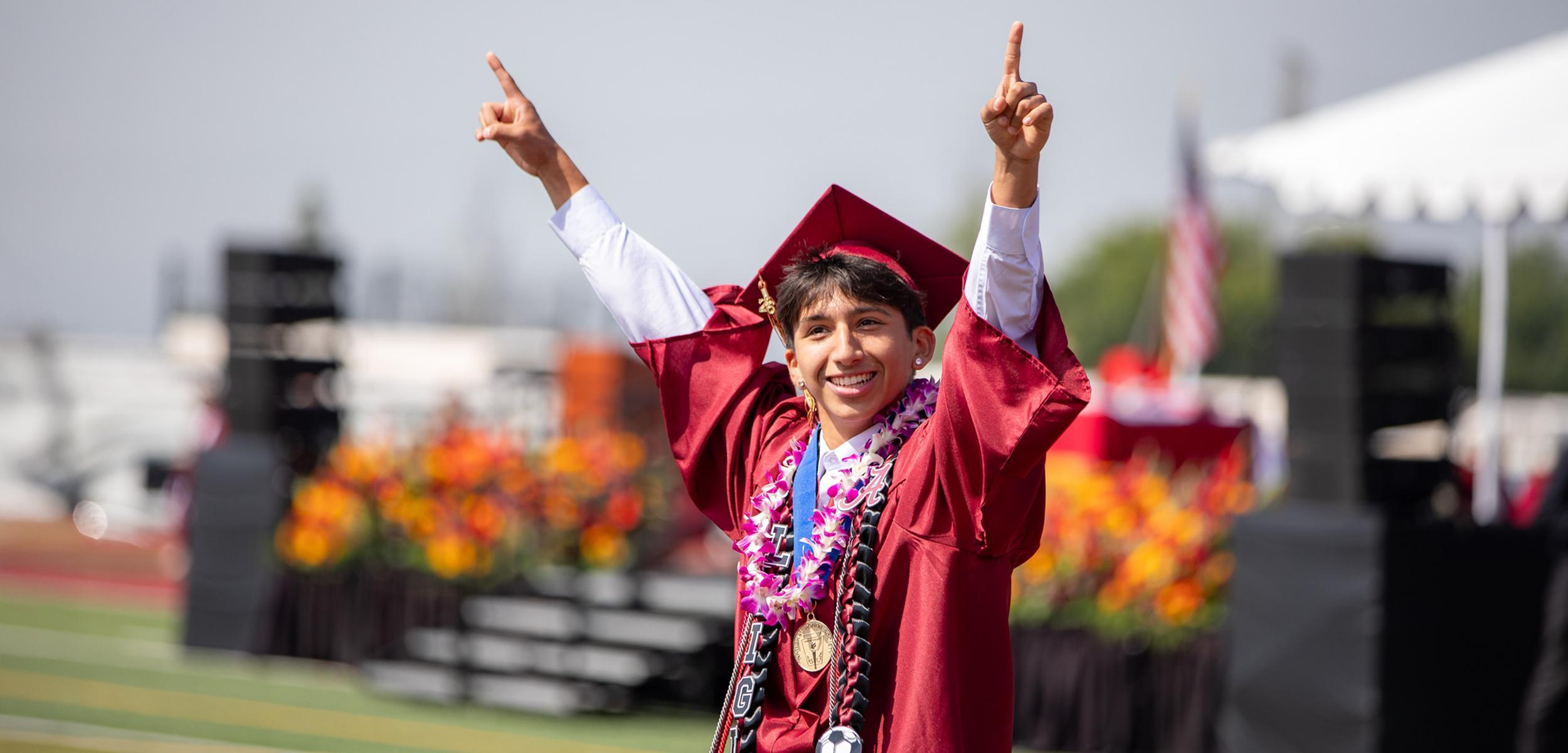 A graduate in a maroon gown celebrates with raised hands, wearing a lei and medals.