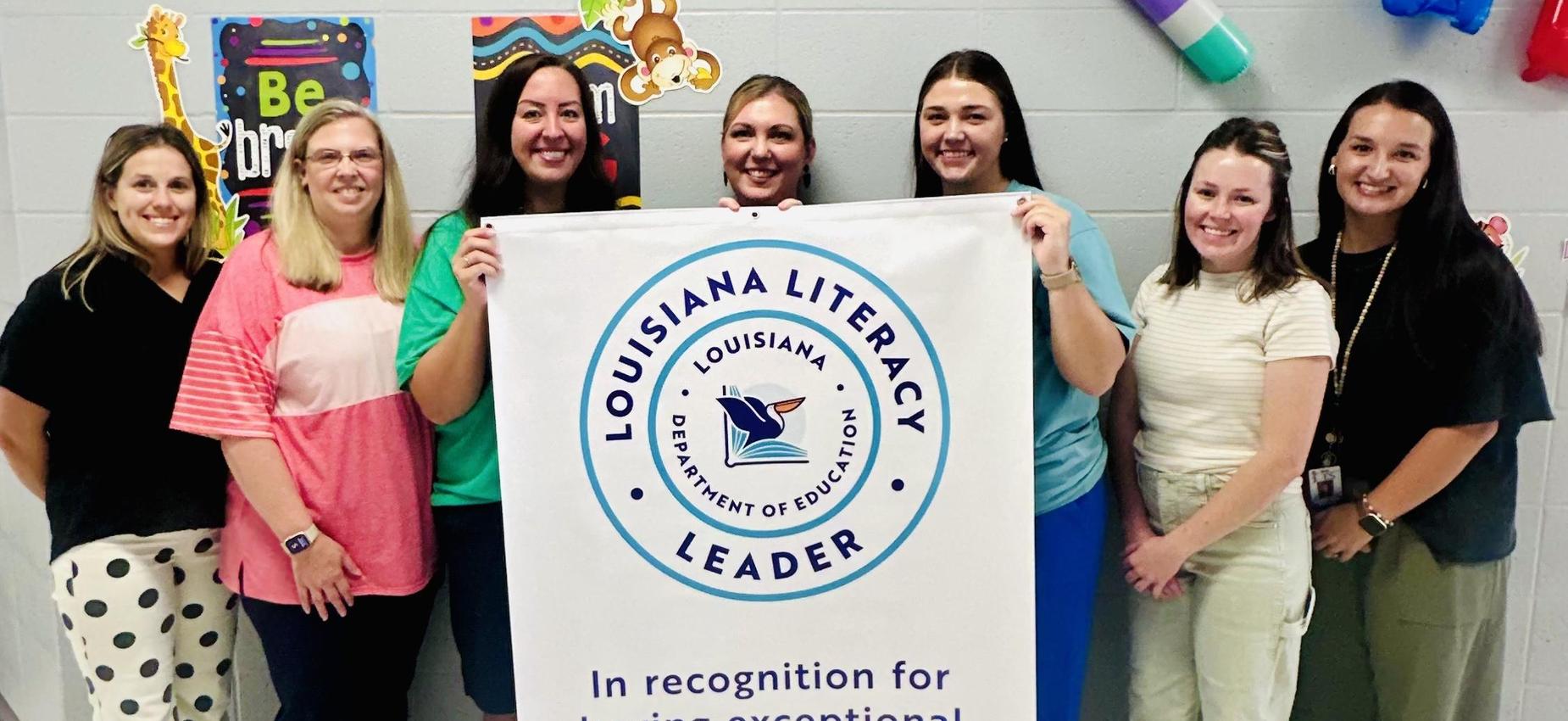 Group of seven women smiling while holding a literacy achievement banner in Louisiana.