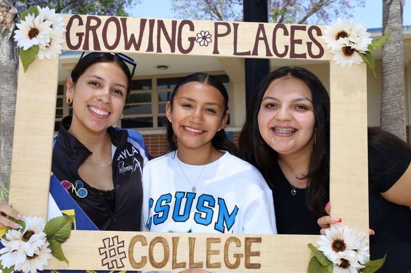 Three students smile behind a decorated “Growing Places” photo frame featuring flowers and a “#College” sign.