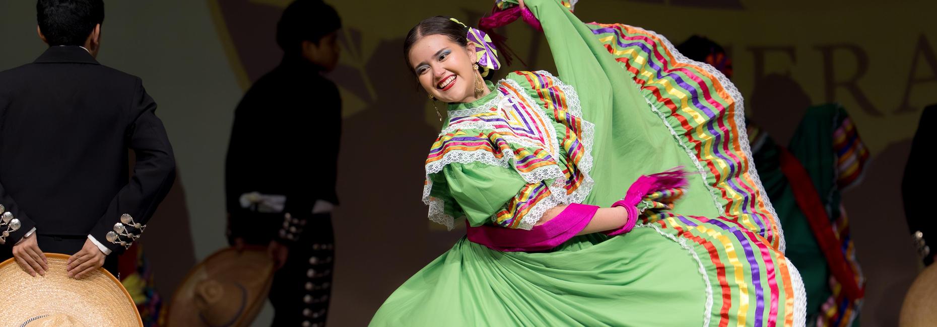 A student performs a traditional folkl&oacute;rico dance on stage while wearing a green dress with colorful ribbon trim.