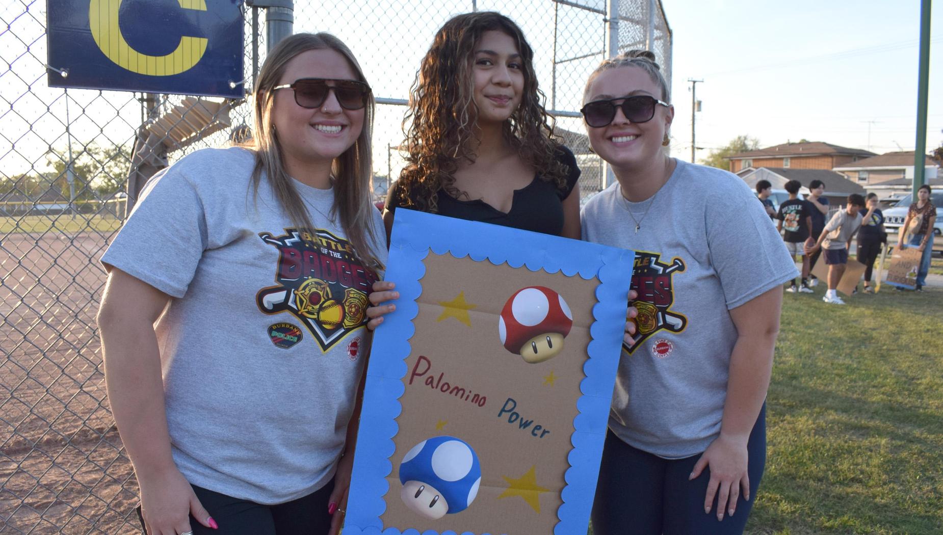 Two women and a girl hold a sign with mushrooms at a softball event.