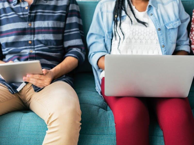 Two people sitting on a couch using a tablet and a laptop.