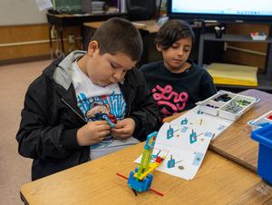 Two young students work together at a classroom table, following step-by-step instructions to build a LEGO Education model. One student focuses intently on assembling a piece while the other observes, surrounded by trays of parts.