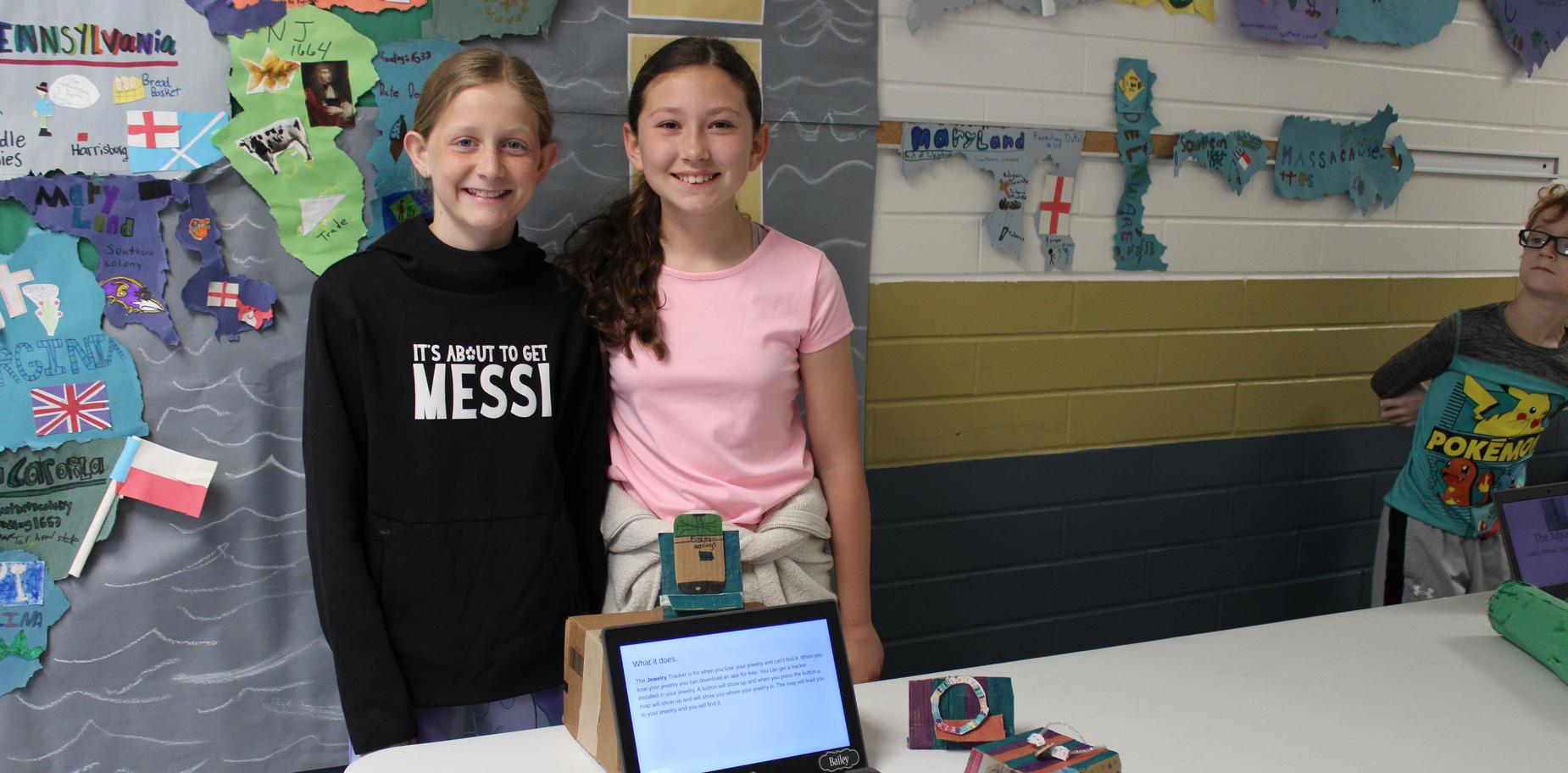 Two girls stand next to each other, showcasing their project at a table.