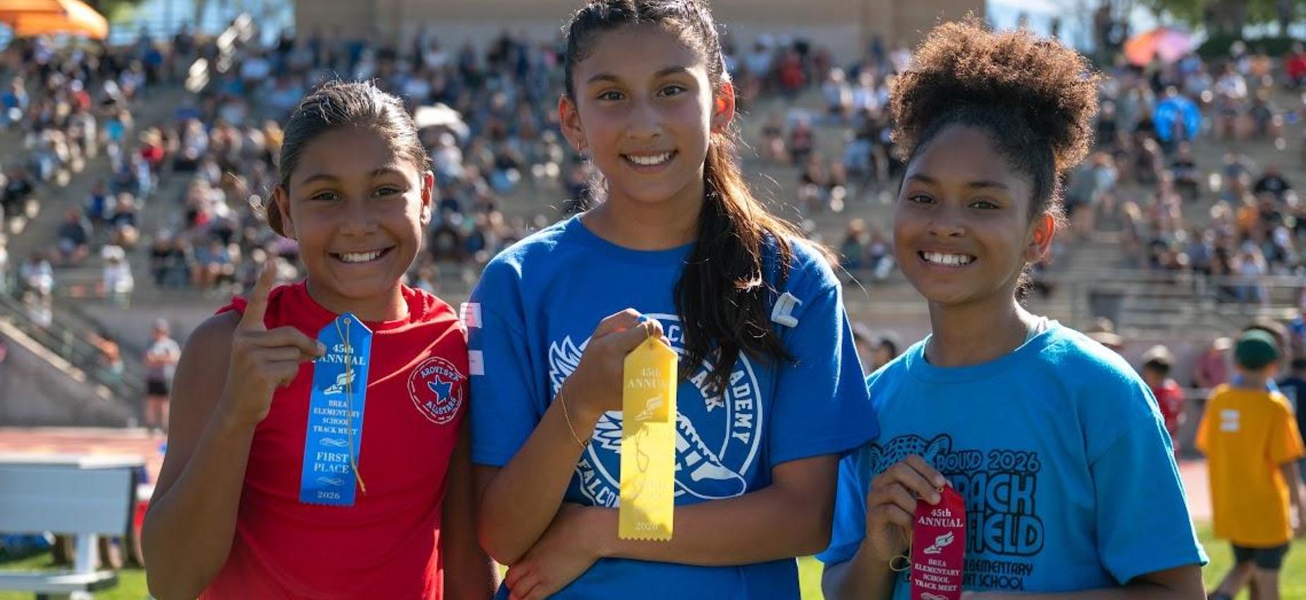 Three smiling girls holding ribbons at a sporting event.