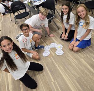 Four children sitting on the floor around a hexagonal game layout, smiling and engaging.