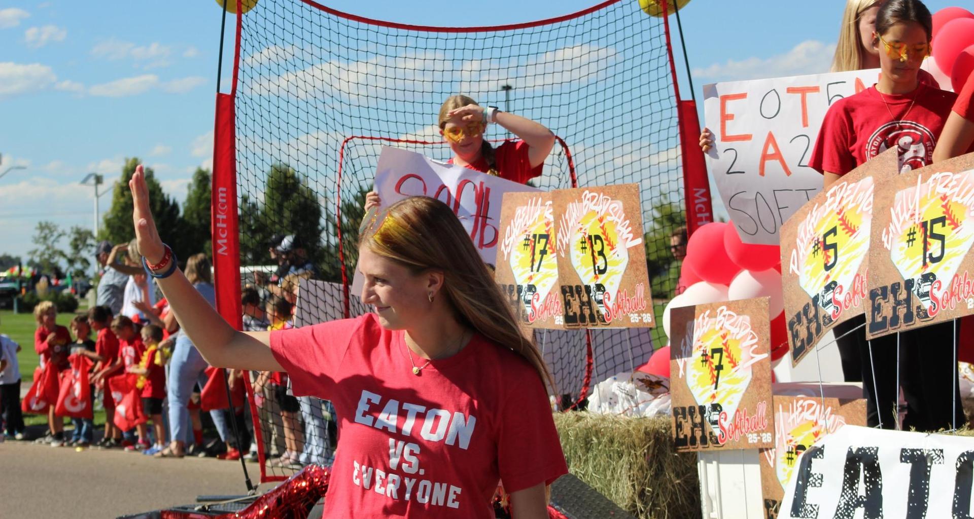 Girl in red shirt waves at a parade with banners and a cheering crowd behind her.
