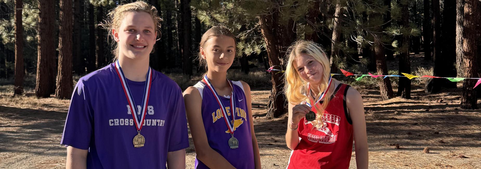 Three young athletes stand together, smiling and wearing medal ribbons in a forest setting.